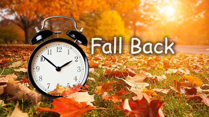 Alarm clock on the ground surrounded by fall leaves with the text Fall Back.