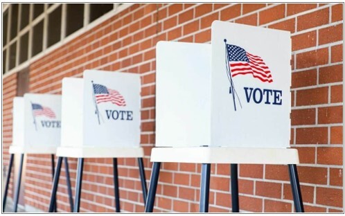 Voting booths lined up against a brick wall with a flag and the text Vote on the side.